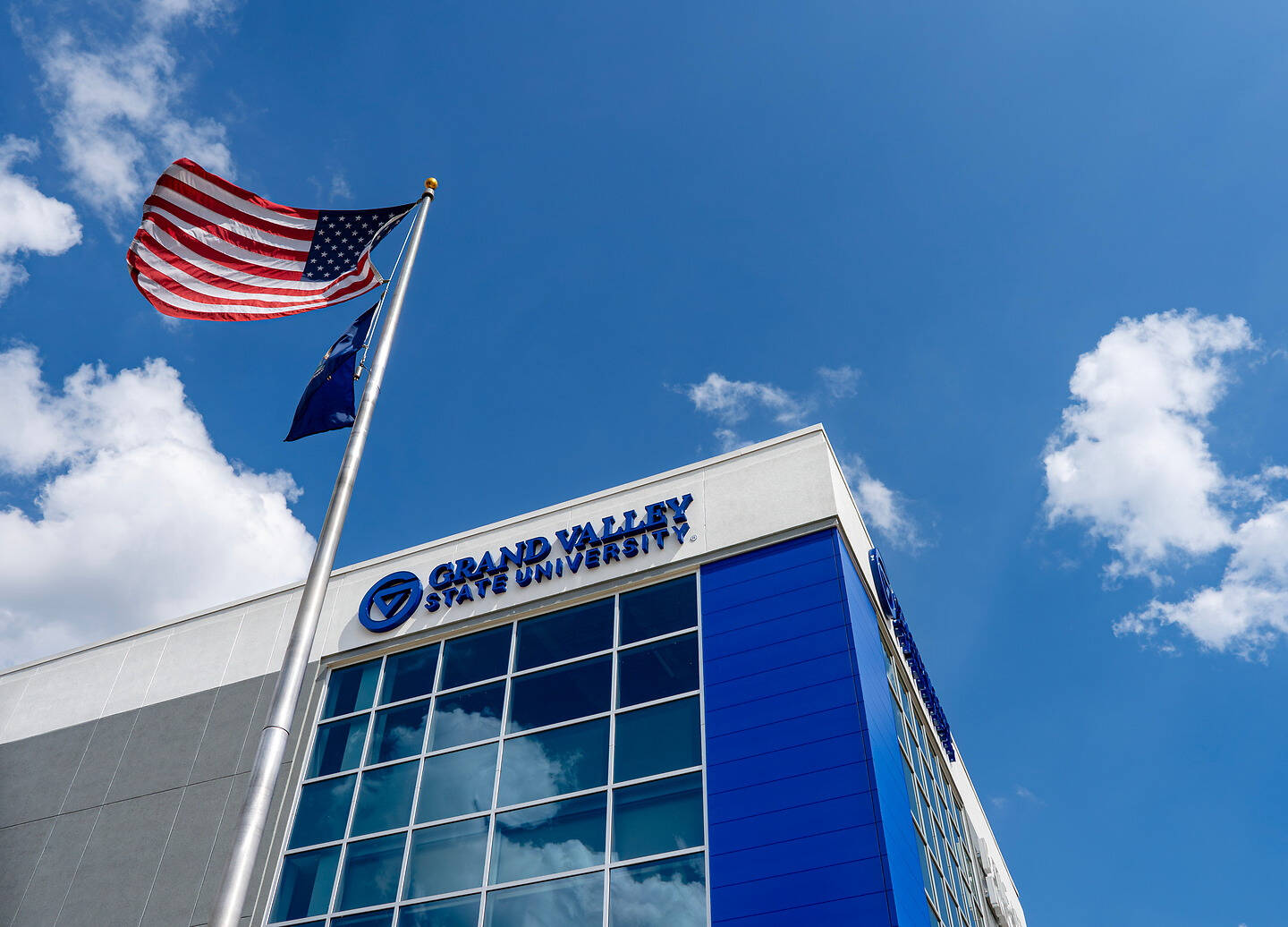An American flag flies beside a modern building with "Grand Valley State University" signage under a bright blue sky.